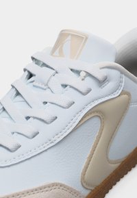 Κοντινό πλάνο λευκού αθλητικού παπουτσιού με μπεζ κυματιστό σχέδιο, λευκά υφασμένα κορδόνια και καφέ λογότυπο στο γλώσσο, που δείχνει λεπτομερή ραφή.