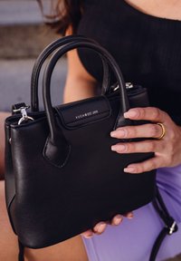 Black leather handbag with rounded handles, silver hardware, and embossed logo. Smooth texture, compact design, held by a hand with neutral nails.