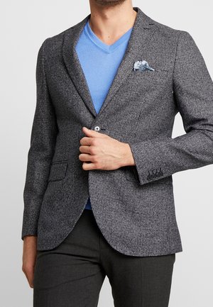 Uomo che sistema un blazer grigio con motivo a quadri a un bottone sopra una maglia a V azzurra chiara e pantaloni scuri, con un fazzoletto floreale nel taschino del blazer.