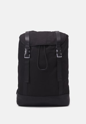 Sac à dos en tissu noir avec rabat supérieur sécurisé par deux sangles noires et boucles argentées, et une base noire renforcée.