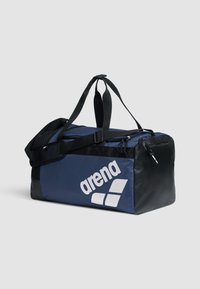 Bolsa de deporte azul con acentos negros, material de tela, un zipper resistente y un logotipo blanco prominente. Cuenta con dos asas y una correa ajustable.