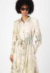 Abito camicia a maniche lunghe in morbido tessuto color crema con stampa di foglie di palma. Presenta un collo alla coreana e una vita aderente con coulisse.