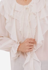 Bílá halenka z lehké látky s výraznými volány vpředu a na rukávech, nastavitelným zavazováním u krku a knoflíkovými detaily.