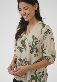 Blusa floreale beige con motivi verdi e blu, scollo a V, maniche corte a palloncino con polsini arricciati, realizzata in tessuto leggero.