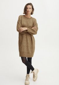 Vestito in maglia marrone con maniche lunghe, vestibilità morbida e scollo a coste; abbinato a collant neri e stivaletti in pizzo beige chiaro.
