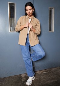 Chemise en velours côtelé marron oversize, débardeur blanc en dessous, jeans baggy bleu clair et baskets blanches. Le décor présente un mur et un sol gris.