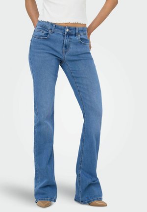 Femme portant un jean bootcut bleu taille mi-haute et des chaussures marron à bout pointu, debout les mains dans les poches arrière, devant un fond uni.