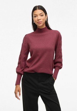 Pull à col montant bordeaux avec des manches ballon texturées, porté avec un pantalon noir. Tissu en maille douce et coupe décontractée.