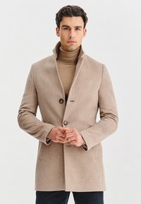 Beżowy płaszcz z mieszanki wełny z wysokim kołnierzem, zapinany na jeden rząd guzików, z dwoma guzikami i bocznymi kieszeniami, noszony na jasnobrązowym golfie.