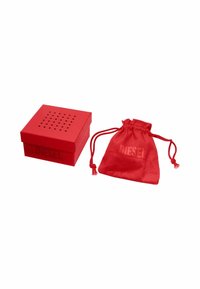 Caja de cartón cuadrada roja con tapa perforada y "DIESEL" en relieve. Al lado, un bolso de satén rojo con cordón de la misma marca.