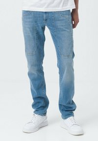 Jeans in denim azzurro chiaro con taglio dritto, toppe rinforzate sulle ginocchia, due tasche anteriori e una texture consumata. Abbinati a sneakers bianche.