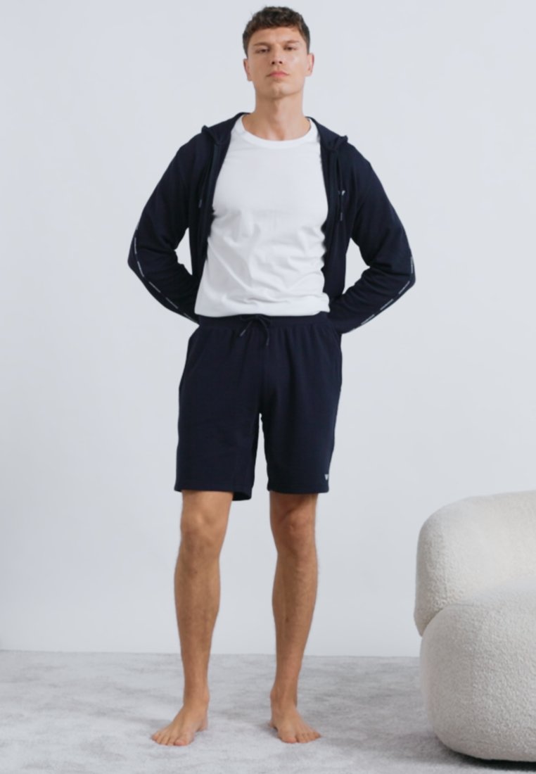 Sweat à capuche zippé marine, T-shirt blanc et short marine. Texture jersey lisse, coupe décontractée, accents sur les coutures latérales, et taille élastique.