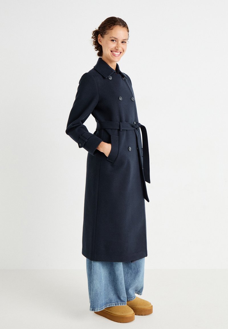Navy double-breasted trench coat with a belt, tailored fit, and button accents, paired with wide-leg denim and brown platform shoes.