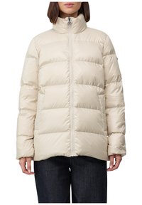 Žena v béžovom prešívanom puffere s vysokým golierom a predným zipsom, skombinovaným s tmavomodrými džínami na bielom pozadí.