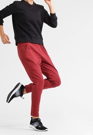 Personne portant un pantalon de jogging rouge et des baskets noires, levant une jambe en plein mouvement, sur un fond blanc uni.