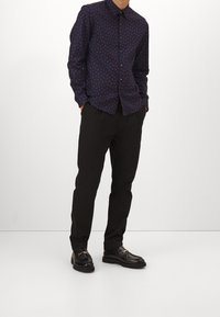 Camicia button-up blu navy con piccolo motivo rosso, abbinata a pantaloni neri. Il modello indossa mocassini neri con dettagli in metallo argentato.