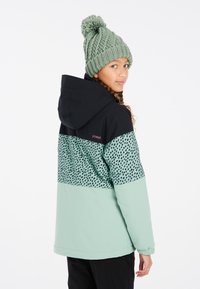 Cappello di maglia verde con pon-pon; giacca nera e menta con motivo a pois nella parte superiore. Texture liscia, vestibilità casual.