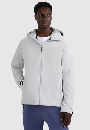 Mann trägt eine hellgraue Kapuzenjacke mit Reißverschluss und die Hände in den Taschen, kombiniert mit dunkelblauen Hosen, vor einem einfarbigen hellen Hintergrund.
