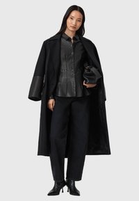 Camicia nera in pelle con chiusura a bottoni, abbinata a un lungo cappotto nero e jeans scuri. Completano il look stivali neri con tacco e una piccola borsetta.