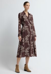 Langes burgunderfarbendes Kleid mit weißem Paisleymuster, Kragen und Knopfdetails an der Taille. Das Modell kombiniert es mit beige-farbenen Stiefeletten. Glattes Material, fließendes Design.