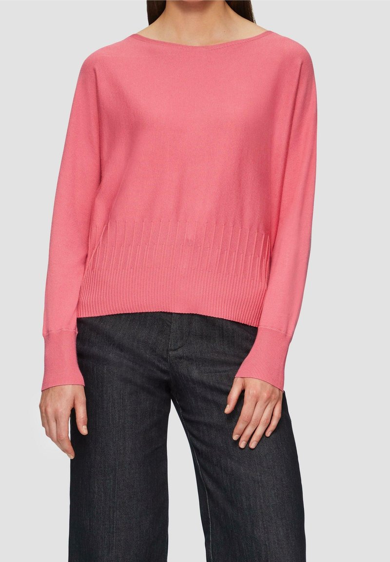 Femme portant un pull rose corail à manches longues avec un ourlet côtelé et un pantalon large gris foncé, debout devant un fond uni.