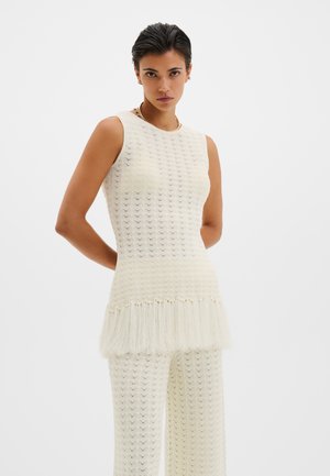 Femme aux cheveux courts et foncés portant un haut tricoté sans manches blanc avec un ourlet à franges et un pantalon large assorti, debout les mains dans le dos.