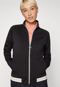 Black zippered jacket with a stand collar, featuring a textured fabric and contrasting striped waistband in beige and white.