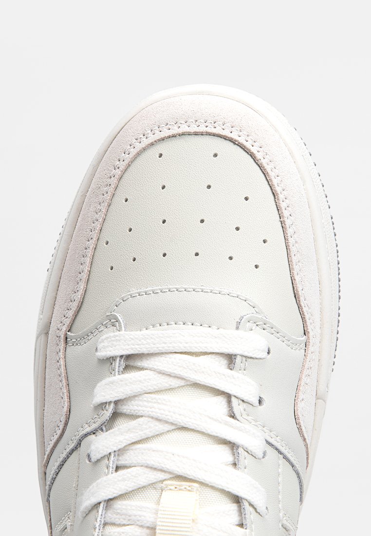 Weißes Leder sneakern mit hellgrauen Wildleder-Akzenten, perforierter Zehenpartie und flachen weißen Schnürsenkeln, mit runder Zehenform und gepolsterter Sohle.