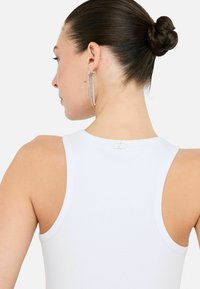 Een vrouw met haar in een knot draagt een witte mouwloze top, waarbij ze hangende oorbellen en een decoratieve sluiting op de rug laat zien.