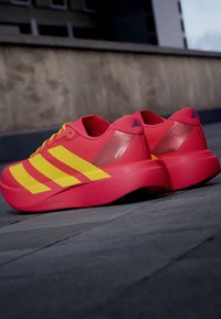 Φωτεινά κόκκινα αθλητικά παπούτσια με τρεις μεγάλες κίτρινες ρίγες και χοντρές σόλες τοποθετημένα έξω σε πεζοδρόμιο.