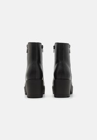 Botas de cuero negro hasta el tobillo con un alto tacón apilado y cremalleras laterales. Textura suave, punta redonda y detalles de costura mínimos.