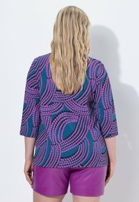 Mehrfarbiges Oberteil mit 3/4-Ärmeln, das einen blauen Hintergrund und pinke sowie schwarze kreisförmige Muster aufweist, kombiniert mit lila Shorts.