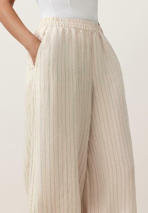 Világos bézs, bő szárú nadrág vékony függőleges csíkokkal, rugalmas derékpánttal és oldalzsebekkel, fehér betűrt felsővel viselve.