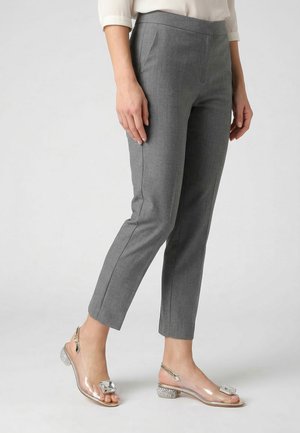 Kobieta w wąskich szarych spodniach i przezroczystych sandałach na niskim obcasie z kwadratową dekoracją z kamieni szlachetnych, stojąca na jednolitym tle.