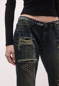 Blugi din denim albastru închis, distressed, cu rupturi, capse metalice și margini destrămate. Prezintă o talie cu capse și patch-uri cu texturi variate.