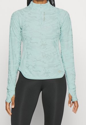 Kobieta ubrana w jasnoniebieską sportową bluzkę z długim rękawem z otworami na kciuki oraz czarne legginsy, stojąca na jednolitym tle.