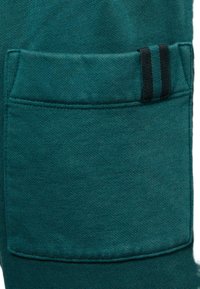 Türkisfarbener Baumwollstoff mit einer großen rechteckigen Tasche, die einen schwarzen und grünen gestreiften Akzent aufweist. Glatte Textur mit gleichmäßigem Gewebe.