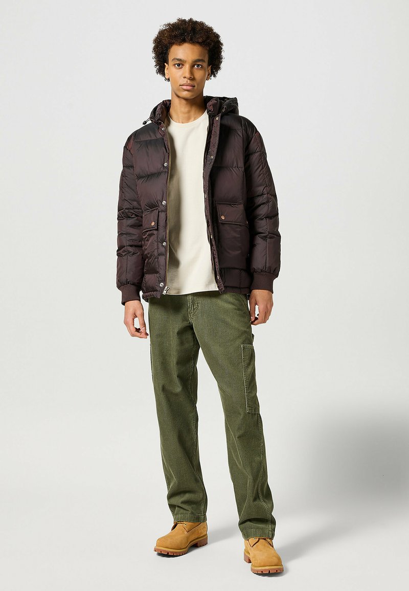 Giubbotto imbottito marrone scuro con cappuccio, dotato di bottoni automatici e tasche laterali. Indossato sopra una camicia beige e abbinato a pantaloni in velluto a coste verde e stivali color beige.