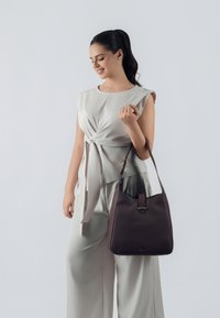 Borsa a tracolla in pelle marrone con una texture liscia, dotata di una sola tracolla e una chiusura superiore. Il modello indossa un top senza maniche grigio chiaro.