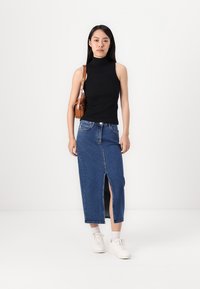 Svart ermeløs topp parret med en blå denimskjørt med en frontspalte. Modellen har på seg hvite sneakers og en liten brun skulderveske.