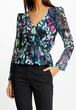 Frau trägt eine langärmelige Blumenbluse mit Rüschen an den Schultern und geraffter Taille, kombiniert mit schwarzen Hosen, Hand in der Tasche.