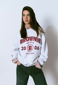 Sudadera gris con "BROWNIE 2006" impreso en burdeos, superpuesta sobre una camisa de collar a rayas, combinada con pantalones verde oscuro.
