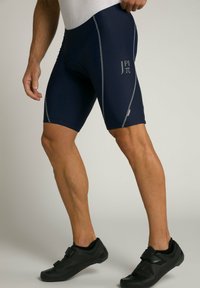 Shorts de cyclisme navy en matériau extensible avec des coutures latérales grises, comportant un logo sur le côté. Portés avec des chaussures de cyclisme noires.