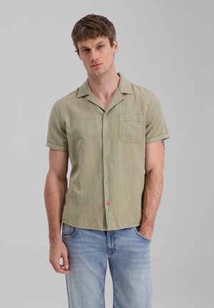 Mann trägt beige kurzärmeliges Hemd mit Brusttasche und hellblaue Jeans, steht vor einfarbigem grauem Hintergrund.