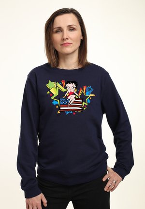 Sweatshirt bleu marine avec un graphique coloré d'une fille de dessin animé assise sur un drapeau américain, entourée d'étoiles vives et d'accents.