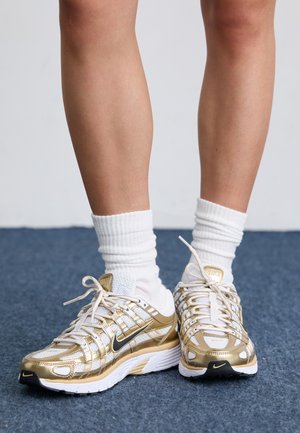 Baskets Nike blanches et dorées avec une tige en maille, des accents métalliques et des détails noirs. Associées à des chaussettes blanches côtelées sur un tapis bleu.