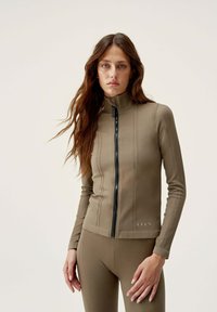 Hnedá zipsová bunda s vysokým golierom, so štruktúrovanými vertikálnymi pruhmi a čiernym zipsom. Spárovaná s rovnakými legínami.