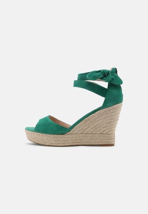Grøn ruskinds kilehæl sandal med åben tå, ankelstropper med en sløjfe og en vævet juteplatform for ekstra højde og tekstur.
