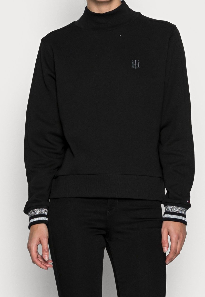 Personne portant un pull à col roulé noir avec des poignets rayés argentés et un petit emblème sur la poitrine, associé à un pantalon noir.