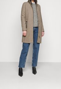 Cappotto in misto lana beige con chiusura con zip, due tasche laterali e taglio dritto. Abbinato a un maglione grigio e jeans blu, con stivali neri.